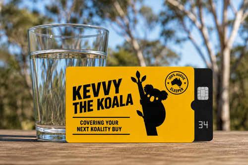 A yellow and black Kevvy The Koala gift card is displayed on a wooden table next to a glass of water, with a blurred outdoor background of trees and blue sky.